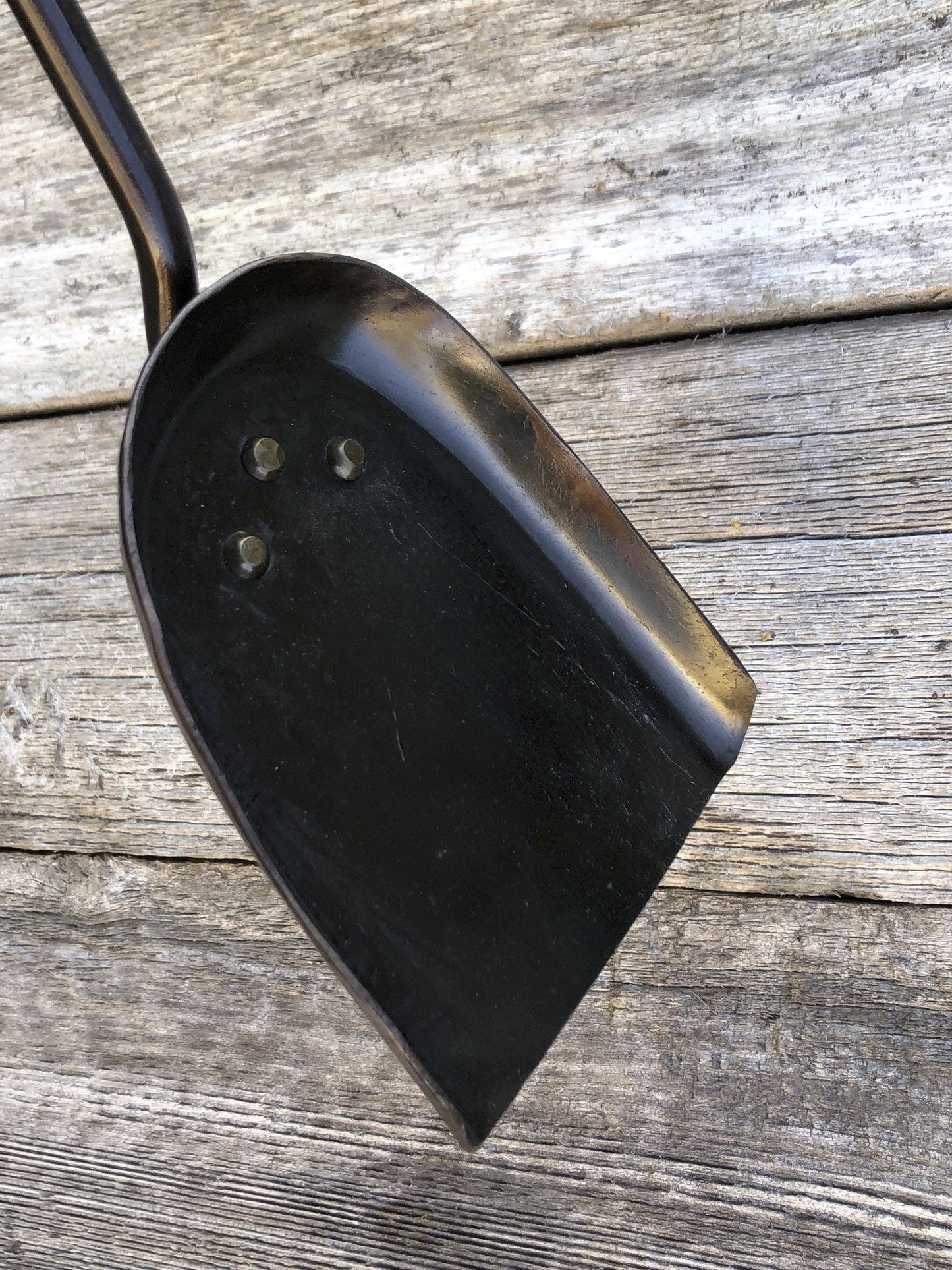 A black hand forged fireplace shovel with a railroad spike handle resting against wooden backdrop.