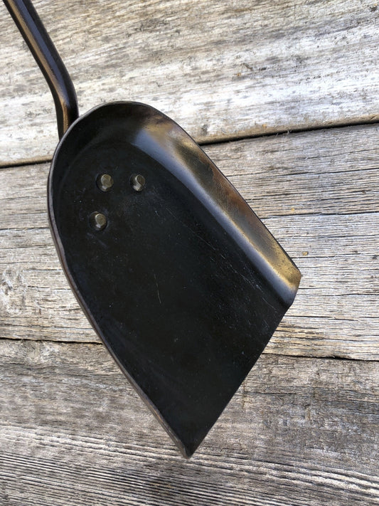 A black hand forged fireplace shovel with a railroad spike handle resting against wooden backdrop.